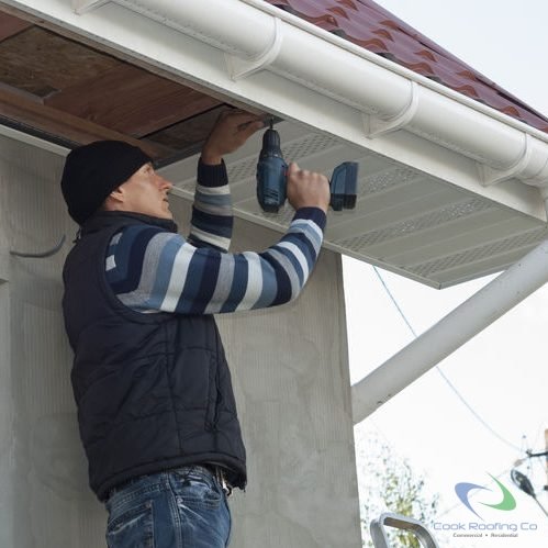 Soffit And Fascia Repair 1 Worker installing vinyl soffit after restoration of extensive storm damage on a stucco house, using power drill. House has upgraded rounded gutters and vinyl fascia