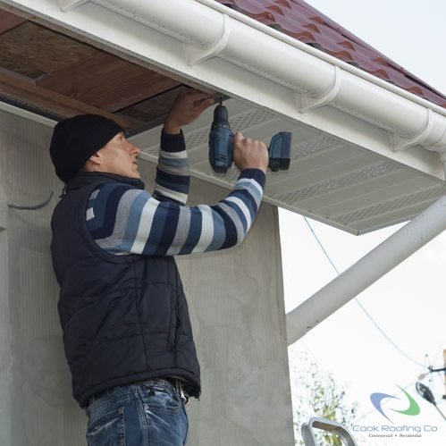 Worker installing vinyl soffit after restoration of extensive storm damage on a stucco house, using power drill. House has upgraded rounded gutters and vinyl fascia